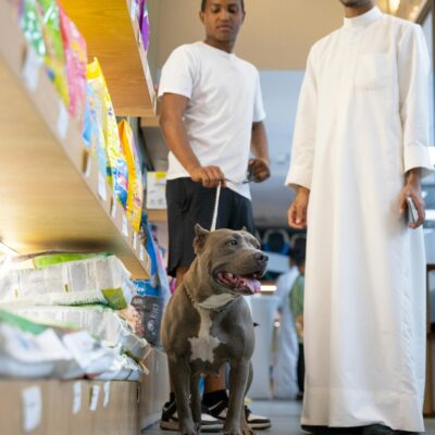 Two men with a pit bull in a pet store aisle exploring dog food options.